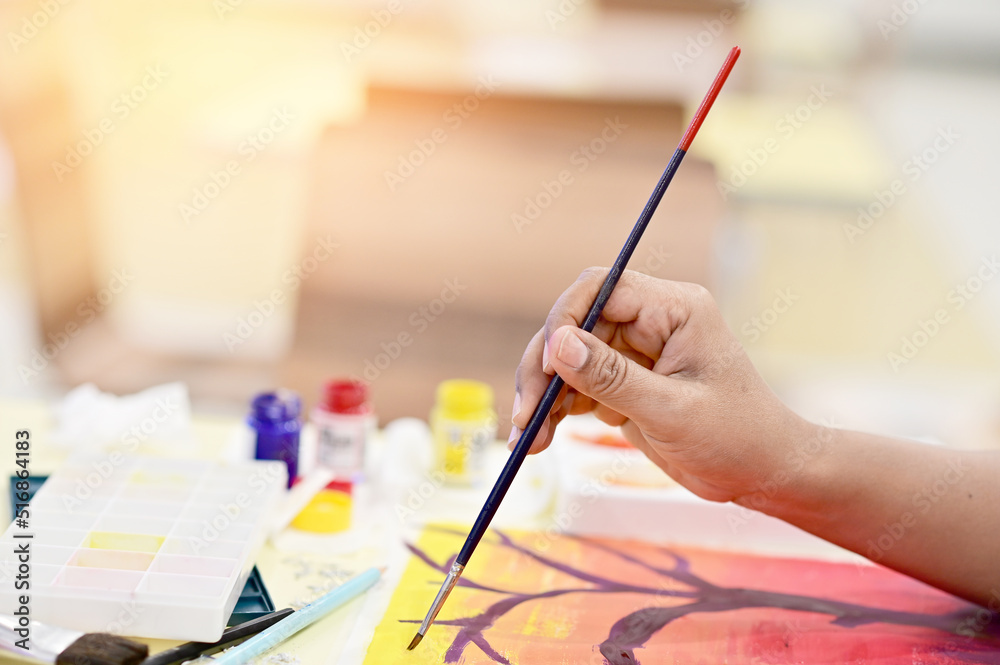 Closeup of Woman hand holding paintbrush with paint bottles and watercolor paintings background. Closeup of Woman hand holding paintbrush with paint bottles and watercolor paintings background.