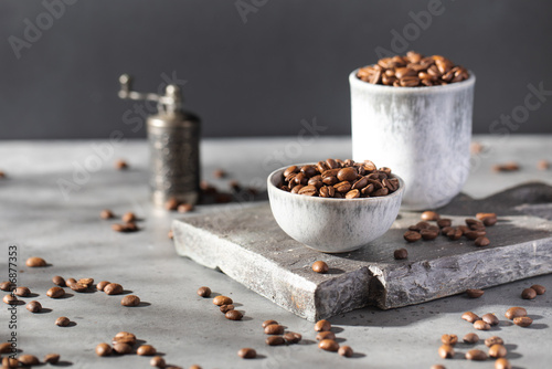 Roasted coffee beans in a bowl on a gray board and coffee grinder on dark background.