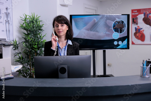 Portrait of smiling hospital receptionist working at front desk using telephone talking with patient making an appointment. Professional looking woman answering phone call at private clinic.