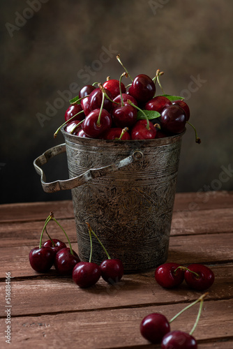 A metal vintage bucket on a wooden table filled with cherries.