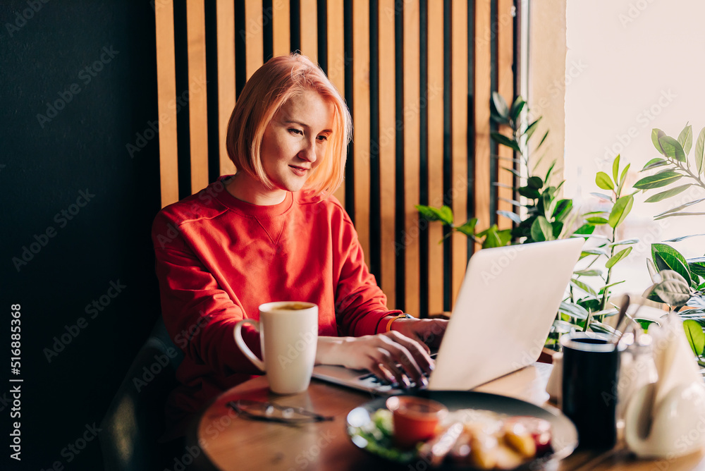 Fototapeta premium Young blonde woman wearing red casual sweater in cafe working on the laptop computer and drinking coffe