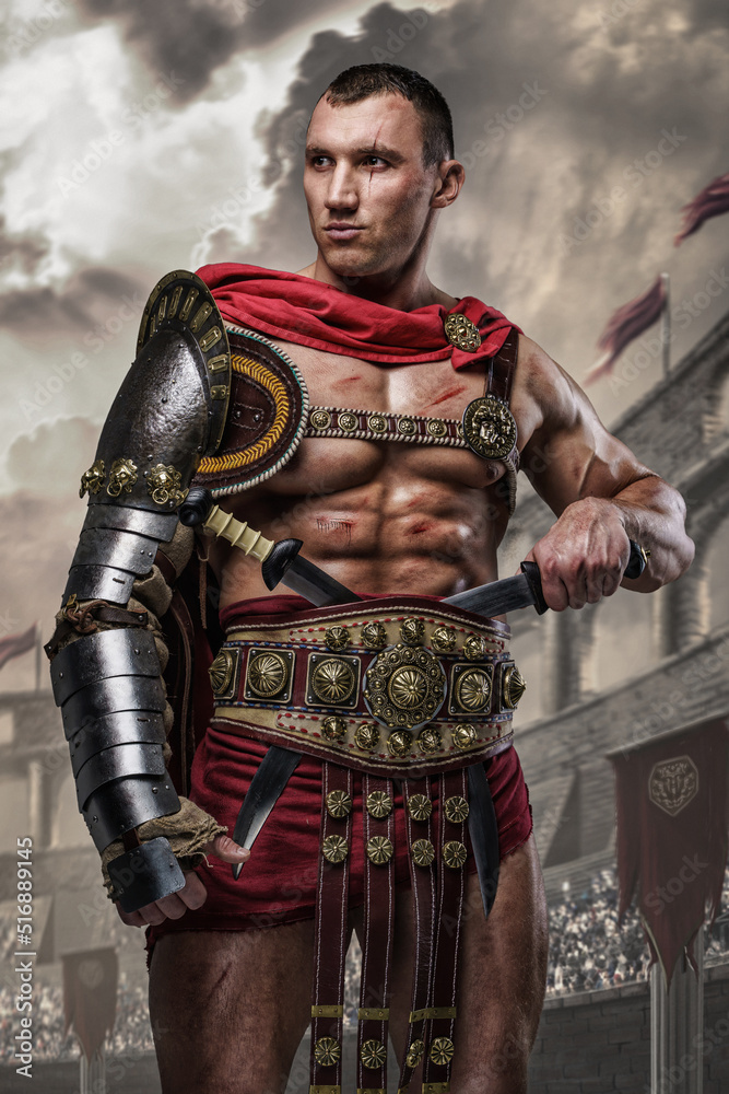 Obraz premium Portrait of handsome gladiator with dual swords dressed in light armor posing in ancient arena.