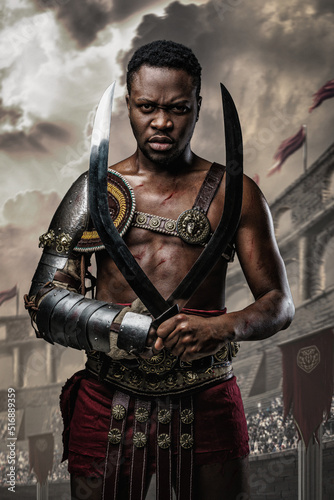 Shot of furious african gladiator holding two swords in cross in arena looking at camera.