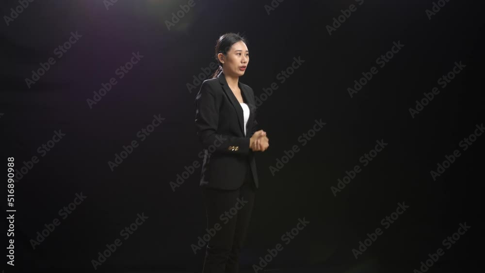 Side View Of Smiling Asian Speaker Woman In Business Suit Showing Thumbs Up Gesture While Standing In The Black Screen Studio
