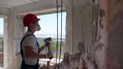 Contractor wrecks wall with sledgehammer making hole for rearrangement. Man doing manual dismantling and demolition works with big hammer hits for apartment renovation. Construction worker in uniform. photo