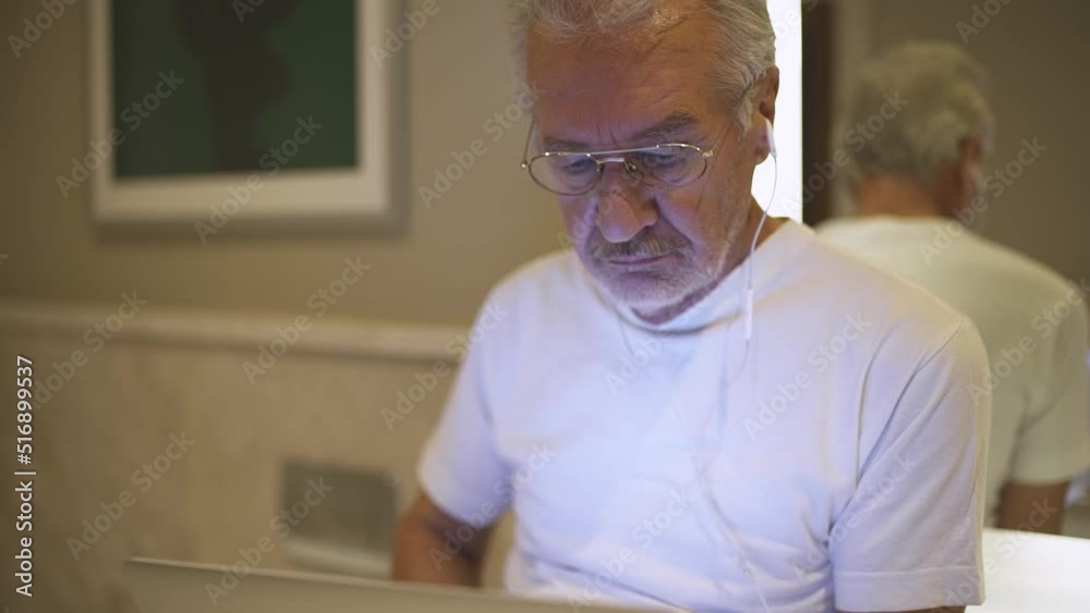 Portrait of an elderly man with glasses looking at a laptop screen, a focused man surfs the internet and listen to music with headphones.