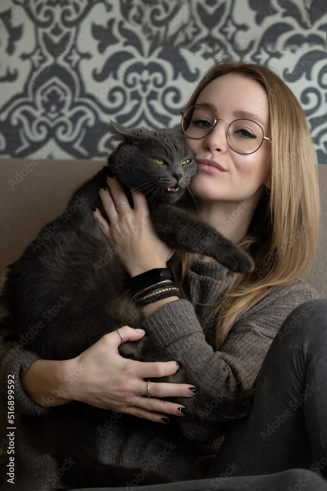 young beautiful woman in glasses at home on the couch holds her gray cat in her arms with a stern character and bright yellow eyes young beautiful woman in glasses at home on the couch holds her gray cat in her arms with a stern character and bright yellow eyes