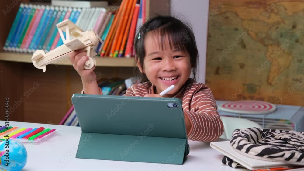 Cute little girl holding a Stylus pen working on a tablet and holding wooden model airplane learning from home with school teacher teaching online. Modern kid and education technology. Cute little girl holding a Stylus pen working on a tablet and holding wooden model airplane learning from home with school teacher teaching online. Modern kid and education technology.