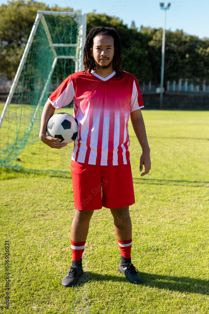 Obraz premium Confident biracial male player with dreadlocks in red jersey holding ball and standing on grass