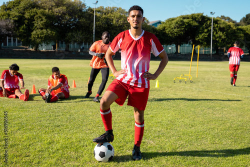 Multiracial confident male player in uniform with arms akimbo and leg on soccer ball at playground photo