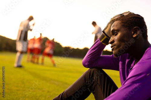 African american stressed male goalkeeper with head in hand sitting on field against clear sky photo