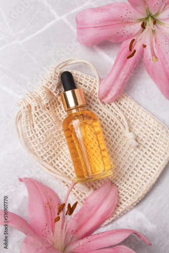 Unbranded serum bottle on cotton bag with lily flower on marble background. Beauty concept for face and body care