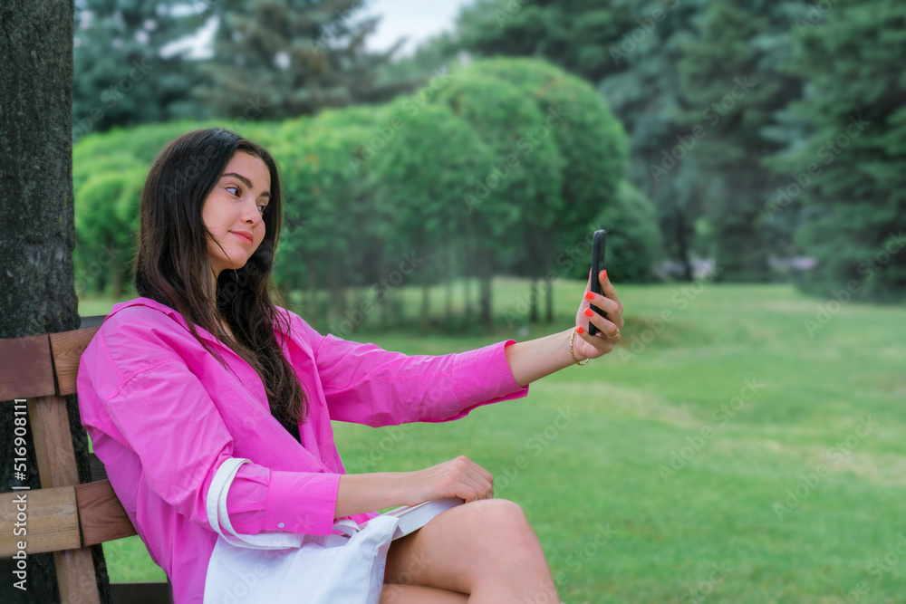 Beautiful teenage girl sitting on wooden bench at city park and taking selfie with smartphone Beautiful teenage girl sitting on wooden bench at city park and taking selfie with smartphone