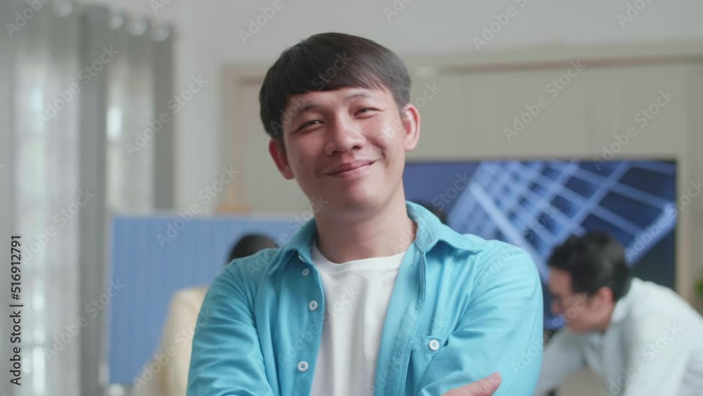 Close Up Of Smiling Asian Man Engineer Crossing His Arms To Camera While His Colleagues Are Discussing About Work Of Solar Cell At The Office
