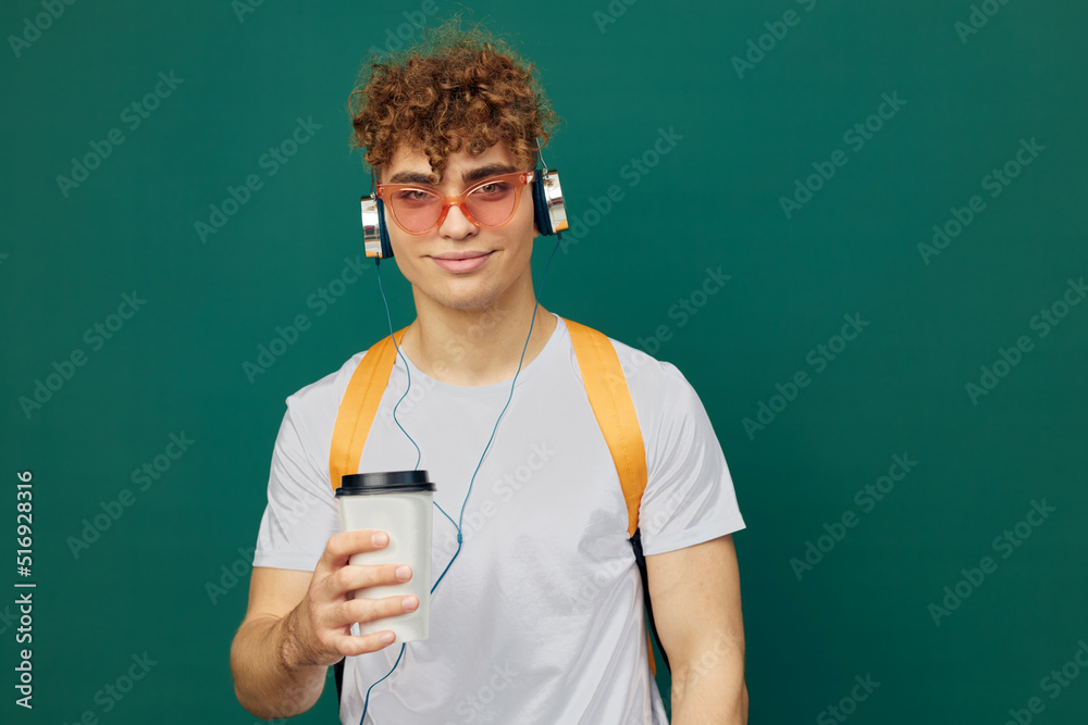 a cheerful man in gray clothes stands on a green background listening to music with headphones with bright glasses and a backpack on his back holding a cardboard glass for hot drinks in his hand a cheerful man in gray clothes stands on a green background listening to music with headphones with bright glasses and a backpack on his back holding a cardboard glass for hot drinks in his hand