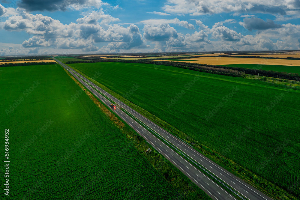 red truck driving on asphalt road along the green fields with a cloudy sky. seen from the air. Aerial view landscape. drone photography. cargo delivery