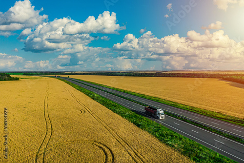 white truck driving on asphalt road along the yellow wheat fields with a cloudy sky. seen from the air. Aerial view landscape. drone photography. cargo delivery © drotik
