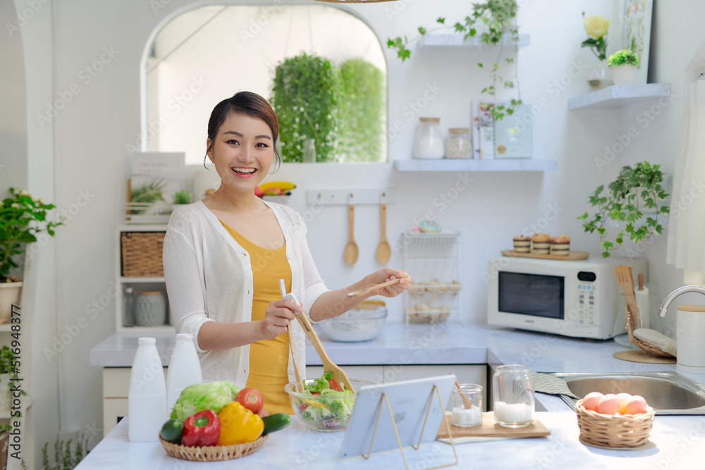 Portrait of smiling young lady cooking fresh organic salad at home in modern kitchen, using and looking at tablet