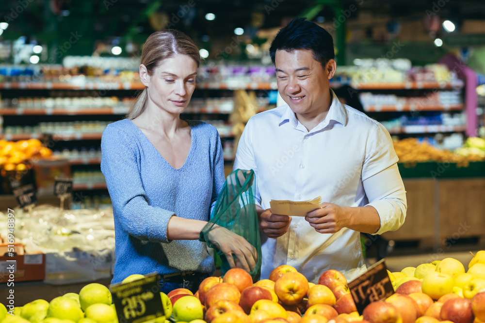 happy mixed race asian couple family man and woman choosing fruits or vegetables in grocery store supermarket. Joint daily shopping together. Buyers customer select product pick an apple in eco bag