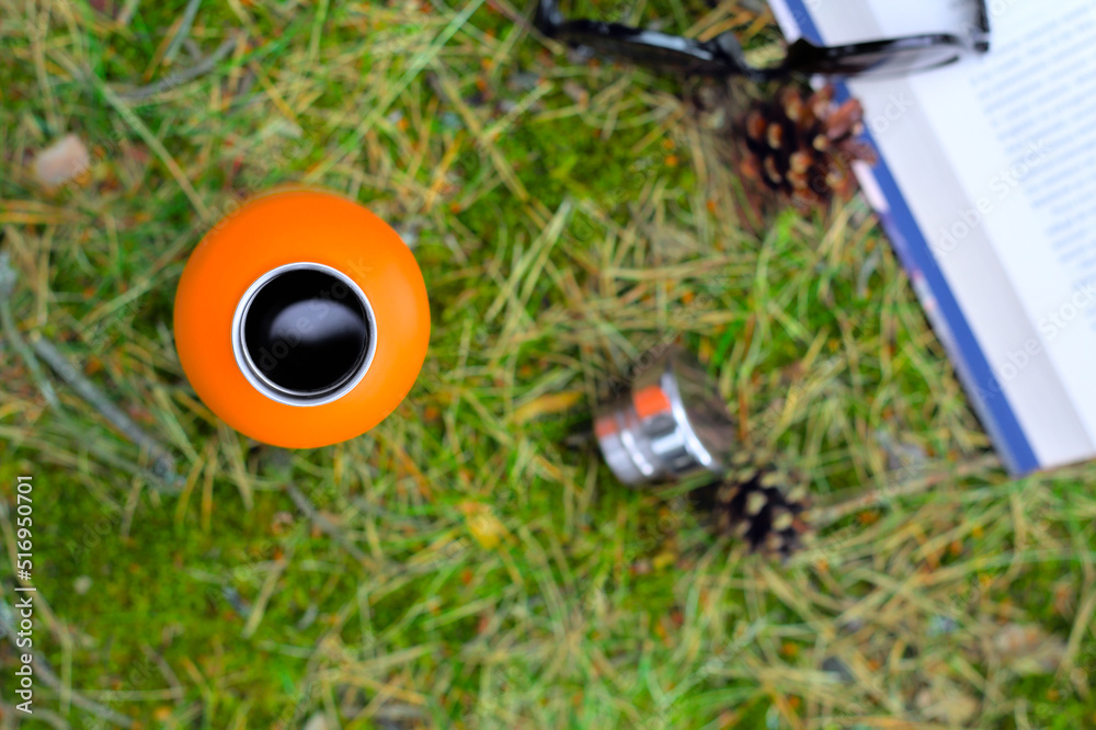 Top view of an orange thermos (bottle) with coffee, a book and sunglasses on a green moss in the forest. A way to relax in nature. Flat lay photo.