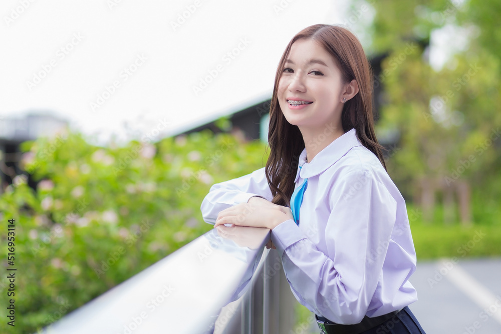 Beautiful high school Asian student girl in the school uniform confidently stands and smiles happily with braces on her teeth while she looks at the camera in a park in the city. Beautiful high school Asian student girl in the school uniform confidently stands and smiles happily with braces on her teeth while she looks at the camera in a park in the city.