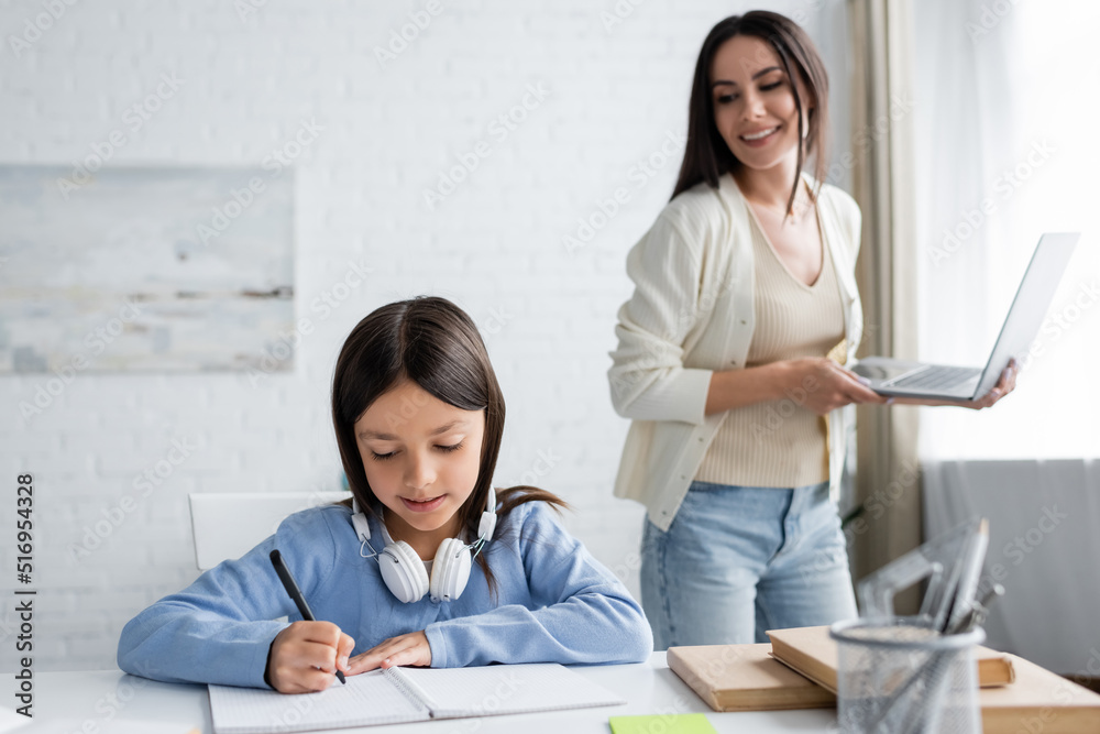 smiling babysitter with laptop near girl writing in notebook at home. smiling babysitter with laptop near girl writing in notebook at home.