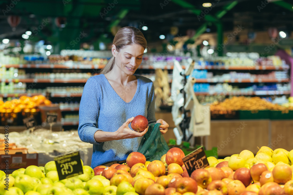 A woman in a supermarket, a buyer chooses an apple fruit, buys and puts an ecological bag in the basket