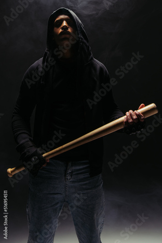 African american bandit in hood holding baseball bat on black background with smoke