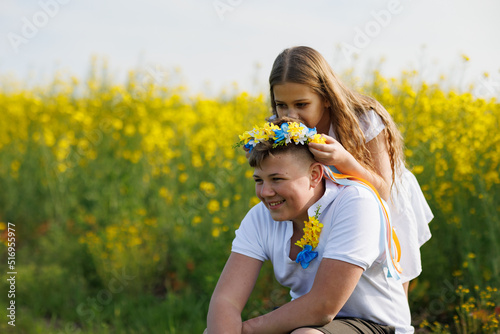 Sister braids ribbons in Ukrainian wreath with flowers on older brother's head, on meadow against field