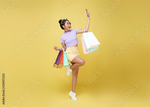 Cheerful happy teen asian woman enjoying shopping, she is carrying shopping bags and smartphone to get the latest offers at the shopping center.