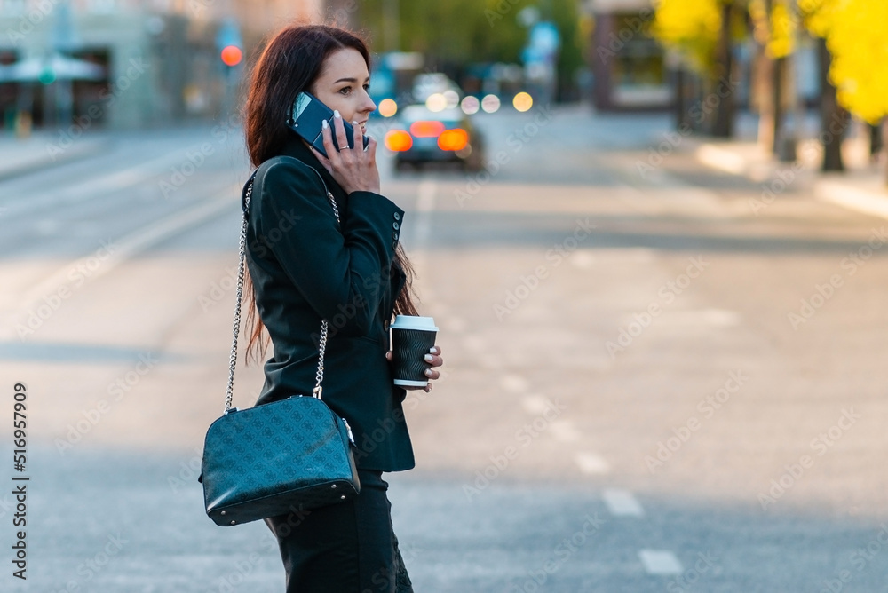 Young business woman talking on the phone holding coffee paper cup,crossing street blurred background. Fashion business photo of beautiful girl in black suite with phone and cup of coffee.Side view.