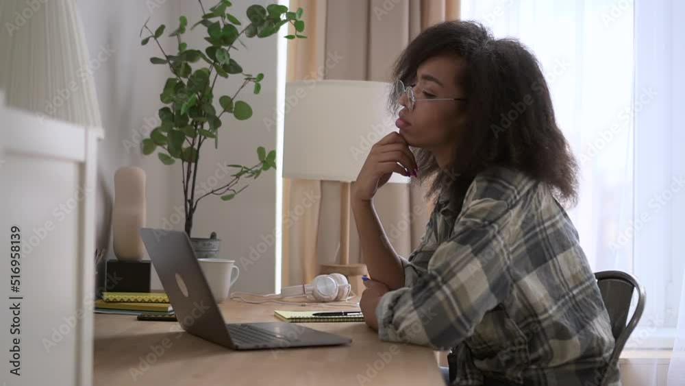 Young woman is typing on laptop and sitting at table in home office spbd. Close view of beautiful American African female thinks about something and looks at computer screen with smile, types text and Young woman is typing on laptop and sitting at table in home office spbd. Close view of beautiful American African female thinks about something and looks at computer screen with smile, types text and