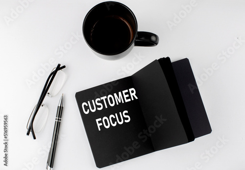 CUSTOMER FOCUS written text in small black notebook with coffee , pen and glasess on white background. Black-white style