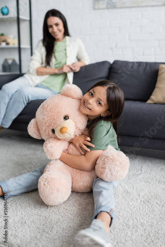 happy girl sitting on carpet and hugging teddy bear near blurred babysitter in living room.
