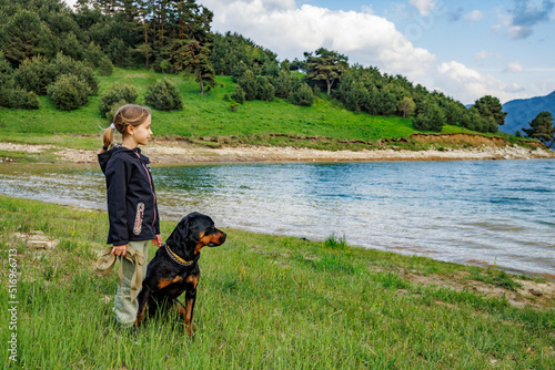 Girl stands near dog of Rottweiler breed in meadow next to lake, against hilly valley with spruce forests © YouraPechkin