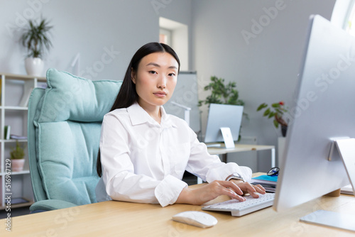 Close-up photo. Portrait of a young beautiful Asian woman secretary sitting at the desk at the computer in the office in business clothes, looking at the camera. © Liubomir