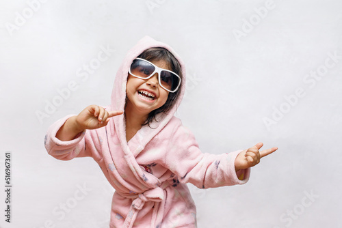 a little brunette girl in retro glasses actively dances with a smile in a pink soft bathrobe with a hood rejoices on a white background, bright, day