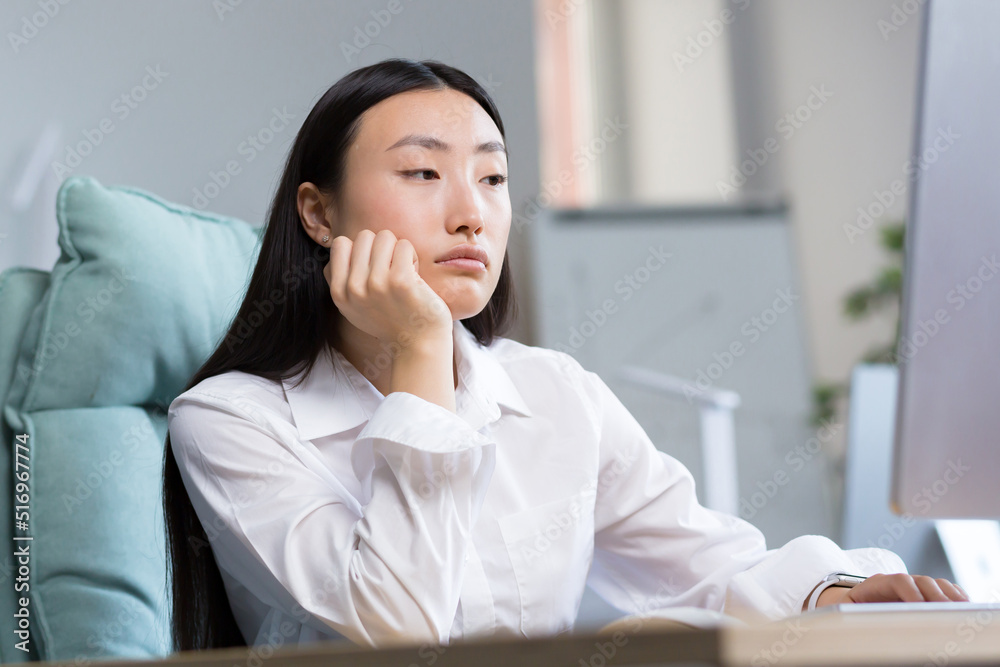 Boring and monotonous work. A young beautiful Asian business woman, an office worker sits at a desk at a computer, is sad, bored, performs monotonous work. He holds his head with one hand. Boring and monotonous work. A young beautiful Asian business woman, an office worker sits at a desk at a computer, is sad, bored, performs monotonous work. He holds his head with one hand.