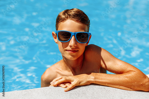 cute boy in sunglasses leaning on the edge of the pool during summer vacation. The boy looks at the camera in the pool