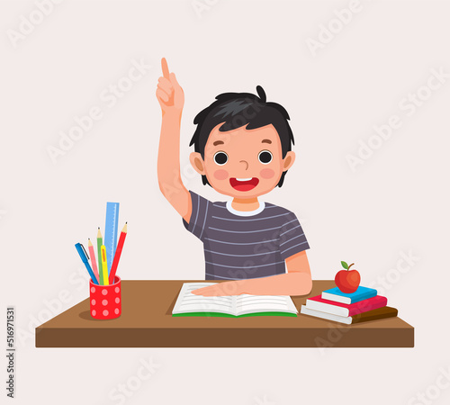 happy little boy student rising hand answering question sitting at her desk in the classroom