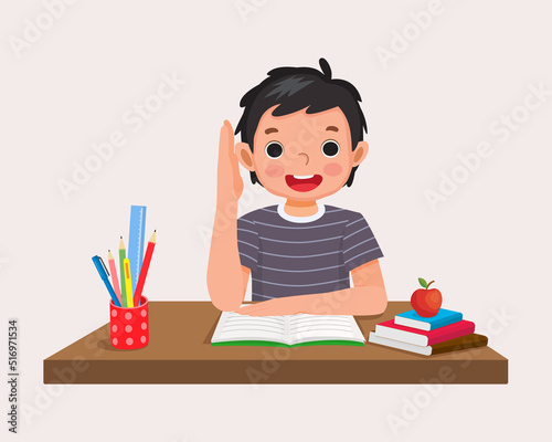 happy little boy student rising hand answering question sitting at her desk in the classroom