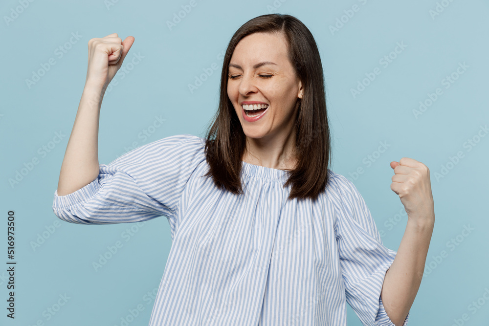 Young happy excited woman she 20s wears casual blouse doing winner gesture celebrate clenching fists say yes isolated on pastel plain light blue background studio portrait. People lifestyle concept. Young happy excited woman she 20s wears casual blouse doing winner gesture celebrate clenching fists say yes isolated on pastel plain light blue background studio portrait. People lifestyle concept.
