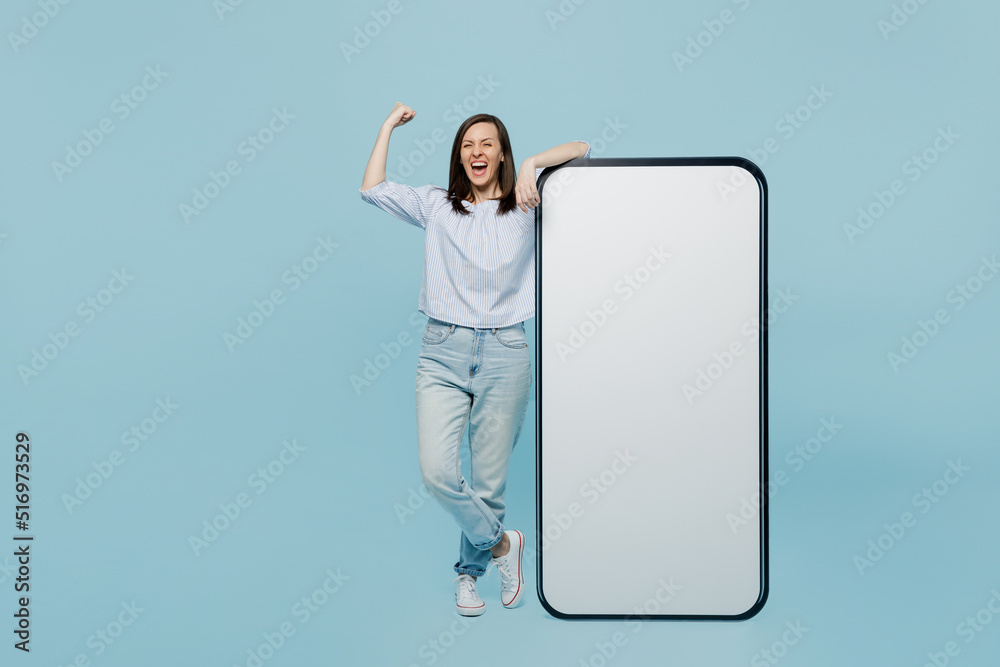 Full body young woman she 20s wear casual blouse big huge blank screen mobile cell phone with workspace copy space mockup area do winner gesture isolated on pastel plain light blue background studio. Full body young woman she 20s wear casual blouse big huge blank screen mobile cell phone with workspace copy space mockup area do winner gesture isolated on pastel plain light blue background studio.