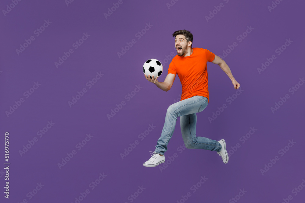 Full size side view excited young fun fan man he in orange t-shirt cheer up support football sport team hold juggling soccer ball watch tv live stream jump run isolated on plain dark purple background