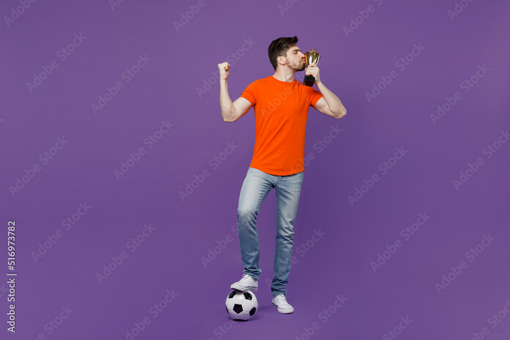 Full body young fan man he wear orange t-shirt cheer up support football sport team hold soccer ball kiss champion cup do winner gesture watch tv live stream isolated on plain dark purple background.