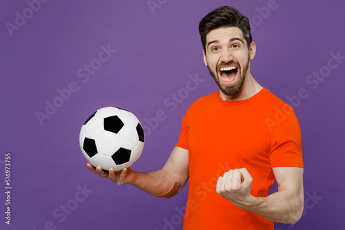 Young fun overjoyed happy fan man he 20s wears orange t-shirt cheer up support football sport team hold in hand soccer ball watch tv live stream do winner gesture isolated on plain purple background.
