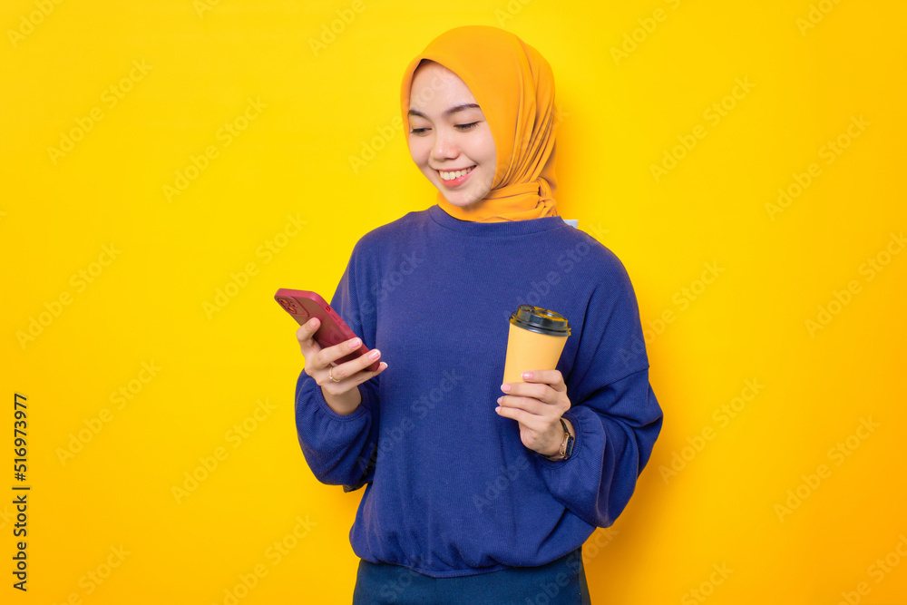 Smiling young Asian Muslim woman dressed in casual sweater using mobile phone for types message, holding coffee cup isolated over yellow background