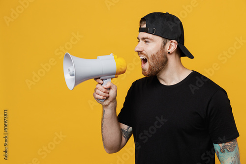 Young bearded tattooed man 20s he wearing casual black t-shirt cap holding scream in megaphone announces discounts sale Hurry up isolated on plain yellow wall background studio. Tattoo translate fun