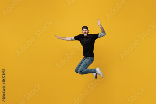 Full body young excited satisfied jubilant bearded tattooed man 20s he wears casual black t-shirt cap jump high with outstretched hands isolated on plain yellow wall background. Tattoo translate fun.