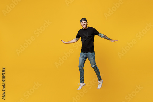 Full body young smiling bearded tattooed man 20s he wears casual black t-shirt cap jump high with outstretched hands isolated on plain yellow wall background studio portrait. People lifestyle concept.